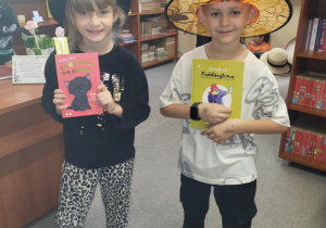 Dwójka uczniów klasy 1 stoi w bibliotece szkolnej podczas pasowania na czytelników. Dziewczynka w czarnym kapeluszu trzyma książkę „Kto przytuli się mną”, a chłopiec w pomarańczowym kapeluszu czarodzieja trzyma książkę „Uściski od Paddingtona”. Oboje uśmiechają się do zdjęcia.