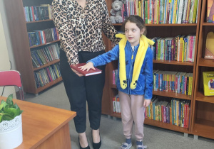 Dziewczynka z klasy 1 stoi obok nauczyciela bibliotekarza i składa przysięgę, trzymając rękę na książce podczas pasowania na czytelnika. W tle widać półki z kolorowymi książkami dla dzieci.