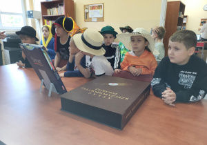 Grupa uczniów klasy 1 siedzi przy stole w bibliotece szkolnej podczas pasowania na czytelników. Dzieci mają na głowach różne kapelusze, a przed nimi leży duży „Atlas Rzeczypospolitej Polskiej” i inne książki. Uczniowie uważnie słuchają przebiegu uroczystości.