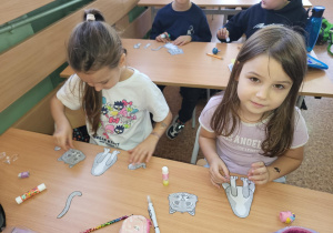 Dwie dziewczynki siedzą przy ławce i składają z papierowych elementów sylwetki kotów podczas zajęć plastycznych w klasie. W tle inni uczniowie również pracują nad swoimi pracami.