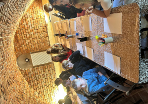 Uczniowie klasy 7a siedzą przy stołach w ceglanej sali i czekają na rozpoczęcie warsztatów plastycznych podczas wizyty w muzeum.