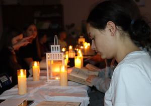 Uczniowie czytają „Dziady” Mickiewicza w przyciemnionej sali oświetlonej świecami, w nastrojowej, skupionej atmosferze.