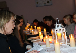Nauczycielka i uczniowie czytają „Dziady” przy świecach i lampionach w nastrojowej, przyciemnionej sali.