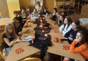 Grupa uczniów klasy 8b siedzi przy dużym stole w szkolnej świetlicy, udekorowanej w halloweenowym stylu. Na stole stoją zapalone świeczki i jedzenie, a uczniowie jedzą i rozmawiają w przyciemnionym nastrojowym oświetleniu.