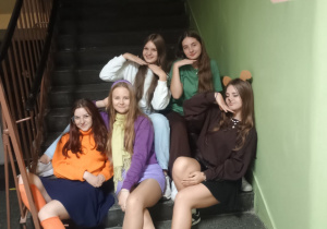 Pięć uczennic klasy 8b siedzi na schodach w szkole podczas nocnej imprezy. Dziewczyny pozują uśmiechnięte, część trzyma dłonie pod brodą w humorystycznym geście. W tle widać poręcz schodów i zieloną ścianę.