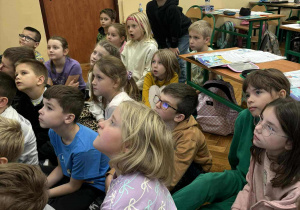 Dzieci w klasie siedzą na podłodze i słuchają czytanego tekstu podczas Sztafety Czytelniczej, patrząc z zaciekawieniem w stronę prowadzącego.