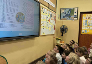Dzieci siedzące na podłodze patrzą na tablicę multimedialną z wyświetlonym tekstem podczas szkolnej Sztafety Czytelniczej.