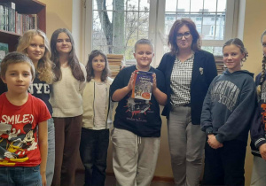 Uczniowie w bibliotece stoją w szeregu obok nauczycielki, a jeden z nich trzyma książkę „Opowieść wigilijna”.