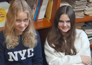 Olga i Ewa z klasy 8b siedzą w bibliotece na tle półek z książkami i uśmiechają się do aparatu.