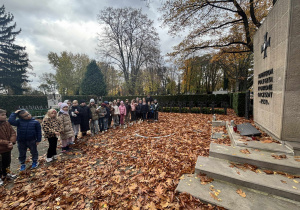 Uczniowie stojący w rzędzie przy pomniku poległych żołnierzy na cmentarzu jesienią, otoczeni opadłymi liśćmi i krzyżami nagrobnymi w tle. Atmosfera pamięci i refleksji.