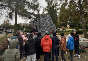 Uczniowie stojący przy pomniku z symbolem Polski Walczącej na cmentarzu, słuchający opiekuna. Jesienna aura, drzewa w tle, uczniowie w kurtkach. Atmosfera pamięci i zadumy.