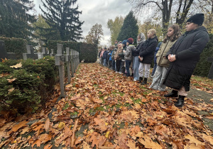 Uczniowie stojący w ciszy przy rzędach grobów żołnierzy na cmentarzu, jesienne liście na ziemi, drzewa w tle. Atmosfera zadumy i pamięci.