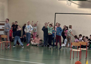 Grupa dzieci na sali gimnastycznej kibicuje podczas zawodów sportowych w świetlicy, w tle stoją krzesła i piłka do koszykówki.