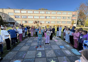 Dzieci ze świetlicy stoją w kręgu na szkolnym boisku wokół rysunków wykonanych kredą; w środku dwie osoby prezentują nagrody podczas zabawy integracyjnej na świeżym powietrzu.