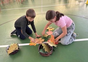 Dwoje dzieci na sali gimnastycznej układa papierowe jesienne liście, biorąc udział w zabawie świetlicowej.