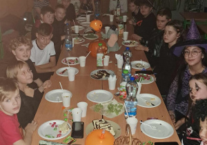 Uczniowie klasy 4a siedzą przy długim stole podczas halloweenowej zabawy w sali lekcyjnej; na stole znajdują się przekąski, napoje i dynie, a dzieci mają różne przebrania i makijaże.