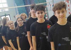 Uczniowie SP 13 w Pabianicach ustawieni w rzędzie na sali gimnastycznej w czarnych koszulkach wolontariatu, stojący przed siatką do siatkówki.