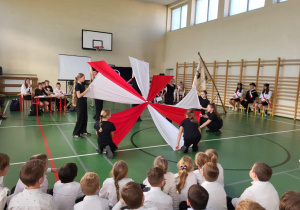 Uczennice prezentują biało-czerwoną rozetę z dużych materiałów podczas apelu niepodległościowego na sali gimnastycznej; na podłodze siedzą młodsi uczniowie, a w tle widać poczet sztandarowy i obserwujących nauczycieli.