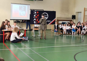 Uczeń w mundurze wojskowym występuje przy mikrofonie podczas apelu niepodległościowego; obok niego uczniowie obsługują prezentację, a w tle widać slajd z Józefem Piłsudskim i dekoracje w barwach narodowych.