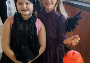 Dwie uczennice klasy 4 w halloweenowych przebraniach stoją na szkolnym korytarzu; jedna jako Wednesday w czarnej sukience i peruce, druga w fioletowej sukience z czarnymi skrzydłami i koszyczkiem w kształcie dyni.