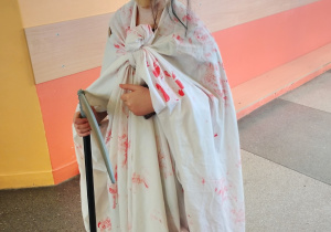 Uczeń klasy 4 w halloweenowym kostiumie z dużą straszną maską i białym prześcieradłem z czerwonymi plamami, trzymająca czarną laskę na szkolnym korytarzu.