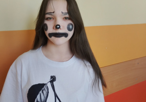 Uczennica klasy 7 w halloweenowym makijażu przypominającym klauna, w białej koszulce z nadrukiem wiśni, stojąca na szkolnym korytarzu.