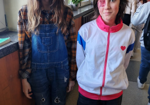 Uczennice klasy 7 stoją na szkolnym korytarzu w halloweenowych przebraniach; jedna w ciemnym makijażu i jeansowym kombinezonie, druga z makijażem imitującym ranę oka w sportowej białej bluzie.