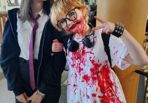 Dwie uczennice klasy 7 w halloweenowych przebraniach stoją na korytarzu; jedna w koszuli z krawatem, druga z mocną charakteryzacją i białą koszulką pokrytą czerwonymi farbami, pozuje z gestem „peace”.