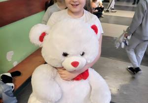 „Dziewczynka uśmiecha się, trzymając dużego białego pluszowego misia z czerwonymi sercami na szkolnym korytarzu.”