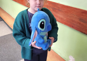 „Uczennica trzyma niebieską pluszową zabawkę podczas obchodów Dnia Pluszowego Misia w szkole.”