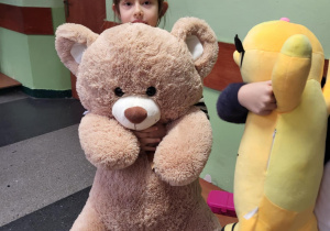 „Dziewczynka przytula wielkiego brązowego pluszowego misia na korytarzu szkolnym.”