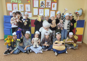 „Uczniowie klasy 1b w opaskach z misiami pozują z pluszakami na tle kolorowych prac plastycznych.”