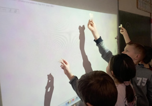 Uczniowie klasy 2b przykładają woskowe kształty do tablicy interaktywnej, obserwując ich cienie w ramach andrzejkowych wróżb.