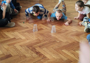 Dzieci z klasy 2a leżą na brzuchu na parkiecie i popychają przedmiot w kierunku trzech plastikowych kubeczków podczas andrzejkowej konkurencji.