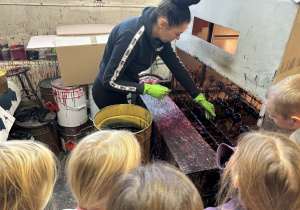 Uczniowie oglądają pracownicę fabryki zanurzającą szklane bombki w kolorowej farbie.