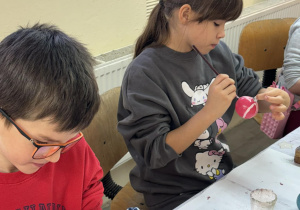 Dwoje uczniów z klas 2a i 2b dokładnie maluje kolorowe bombki pędzelkami przy stole z farbami.