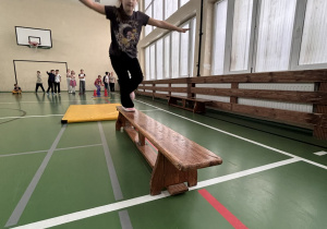 Dziewczynka biegnie po drewnianej ławce na sali gimnastycznej podczas świetlicowej olimpiady, a jej koledzy obserwują konkurencję w tle.