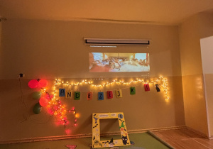 Dekoracja andrzejkowa w świetlicy – napis „Andrzejki”, światełka, balony i żółta fotoramka ustawione pod ekranem, tworzące miejsce do zdjęć dzieci.