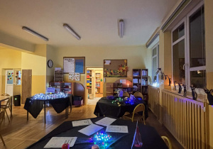 Pusta świetlica udekorowana na andrzejki, stoły z czarnymi obrusami, kartami z wróżbami, kostkami i kolorowymi lampkami czekają na przyjście dzieci.