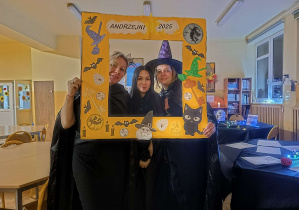 Trzy nauczycielki w andrzejkowych przebraniach pozują w żółtej fotoramce „Andrzejki 2025” w udekorowanej świetlicy.