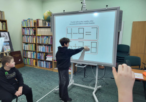 Chłopiec z klasy 1b stoi przy tablicy interaktywnej i wskazuje poprawne przestawienie zapałki w działaniu matematycznym.