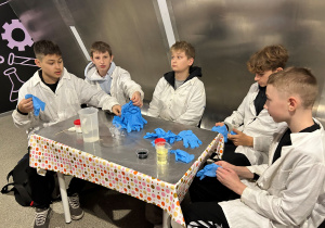 Uczniowie w fartuchach przy stoliku w Experymentarium szykują rękawiczki i odczynniki do doświadczeń na warsztatach „Naukowy misz-masz”.