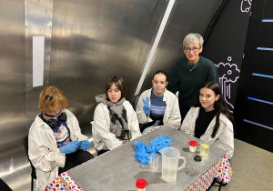 Uczennice klasy 7a w fartuchach i nauczycielka pozują przy stole z odczynnikami podczas warsztatów chemiczno-fizycznych w Experymentarium.