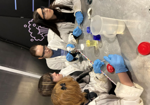 Uczennice klasy 7a w fartuchach i okularach ochronnych wykonują doświadczenie z kolorowymi roztworami podczas warsztatów ‘Naukowy misz-masz’ w Experymentarium.