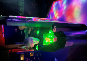 Uczeń klasy 7a na arenie Laser Games trzyma podświetlony na zielono pistolet laserowy, przygotowany do oddania kolejnego strzału.