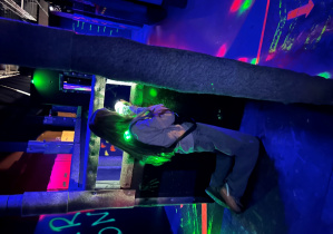 Uczennica 7a klęczy za osłoną na arenie Laser Games i mierzy z pistoletu laserowego w półmroku z kolorowymi światłami.