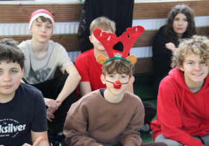 Uczniowie w świątecznych dodatkach siedzą na sali gimnastycznej podczas Mikołajek.