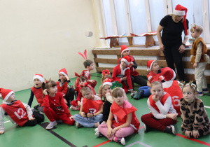 Dzieci w mikołajkowych strojach siedzą na sali gimnastycznej; obok nauczycielka SP 13.