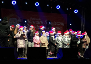 Grupa dzieci w zimowych kurtkach i czapkach Mikołaja stoi na oświetlonej scenie Pabianickiego Jarmarku Świątecznego, przygotowana do świątecznego występu.