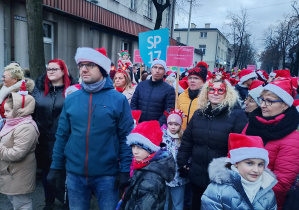 Tłum dzieci i dorosłych w czerwonych czapkach Mikołaja maszeruje ulicą podczas Pabianickiej Parady Świątecznej, w środku widoczny szyld Szkoła Podstawowa nr 13.